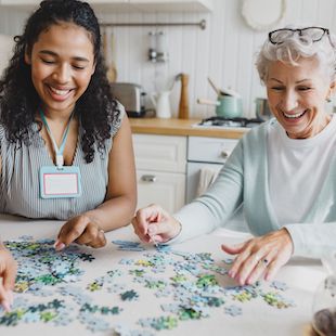 Portrait of african american female social worker volunteer playing puzzle game with cheerful senior lady, having fun, laughing searching for right piece sitting together at kitchen table Portrait of african american female social worker volunteer playing puzzle game with cheerful senior lady, having fun, laughing searching for right piece sitting together at kitchen table